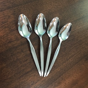 May include: Four silver spoons with rounded, slightly pointed ends. The spoons are arranged in a fan shape on a dark brown wooden surface.