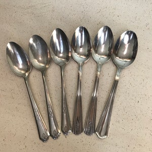 May include: Six silver spoons with ornate handles. The spoons are arranged in a semi-circle on a light beige surface.