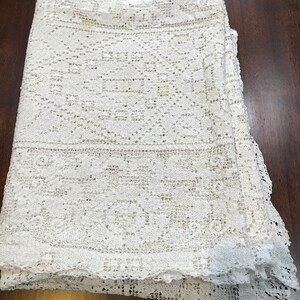 May include: A white crocheted tablecloth with a delicate floral pattern. The tablecloth is folded in thirds and has a scalloped edge.
