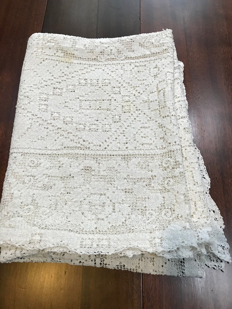 May include: A white crocheted tablecloth with a delicate floral pattern. The tablecloth is folded in thirds and has a scalloped edge.