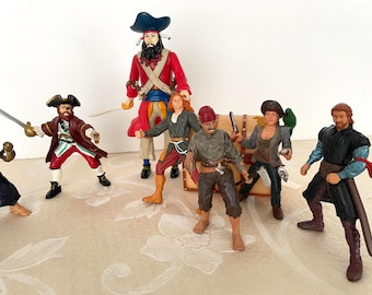 May include: A group of toy pirate figures. The figures are dressed in various pirate outfits, including red coats, hats, and swords. A treasure chest is also present. The figures are posed in action.