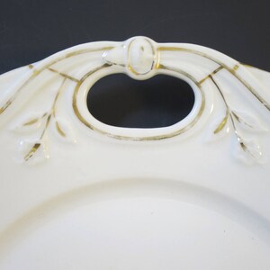 Puede incluir: Plato de servir de porcelana blanca con un borde dorado y un asa decorativa. El asa tiene forma de lazo con un ribete dorado y un peque&ntilde;o pomo en el centro.