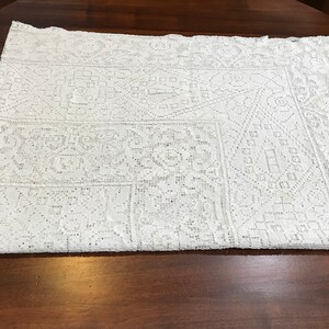 May include: White crocheted tablecloth with a geometric pattern. The tablecloth is folded in half and laid on a wooden surface.