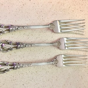 Peut inclure: Trois fourchettes en argent avec des manches ornés d'un motif floral. Les fourchettes sont disposées en rangée, les manches pointant vers la gauche et les dents vers la droite.