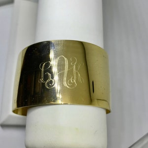 May include: A gold-toned metal cuff bracelet with the monogram "BAK" engraved on the front.