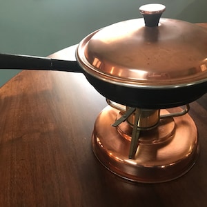 May include: Copper-colored fondue pot with a black handle and lid. The pot sits on a copper-colored stand with a brass-colored burner. The pot is on a dark brown wooden table.