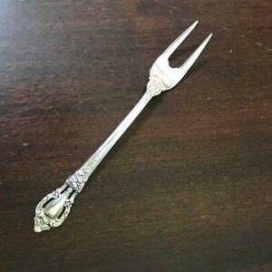 May include: A silver fork with an ornate handle and two prongs. The fork is on a dark brown wooden surface.