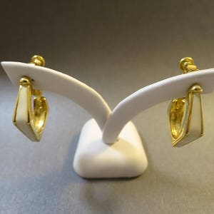 May include: A pair of gold hoop earrings with white enamel accents. The earrings are displayed on a white stand.