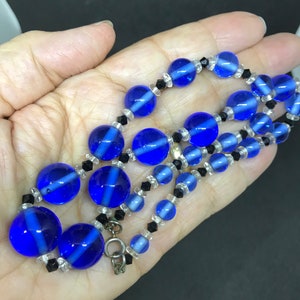 May include: A vintage necklace with large, round, cobalt blue glass beads with white stripes, and smaller clear and black beads. The necklace is shown on a hand, highlighting its design and colour.