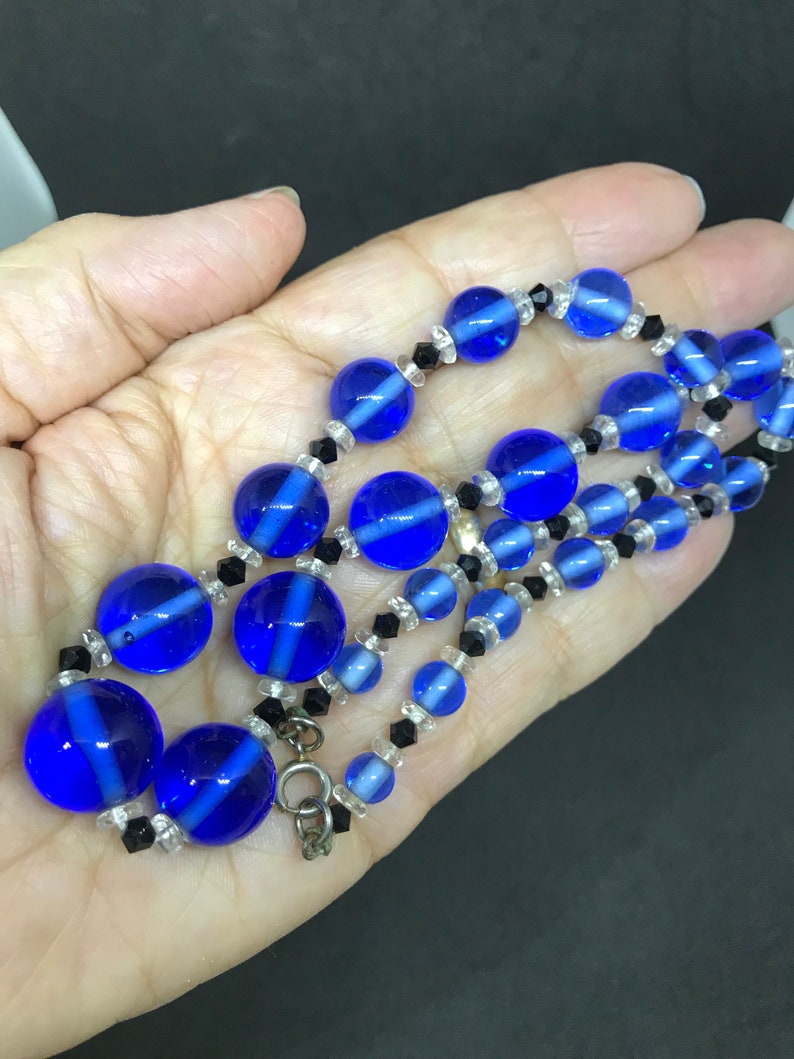 May include: A vintage necklace with large, round, cobalt blue glass beads with white stripes, and smaller clear and black beads. The necklace is shown on a hand, highlighting its design and colour.