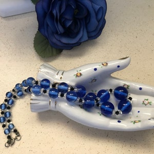 May include: A blue beaded necklace with clear and black accents is displayed on a white ceramic hand. A blue fabric flower is in the background. The necklace features large, round blue beads and smaller black beads.