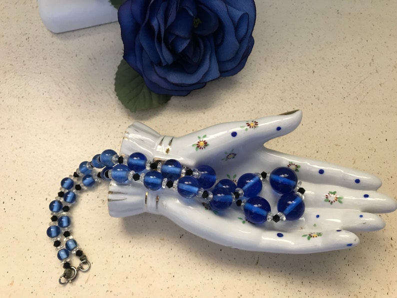 May include: A blue beaded necklace with clear and black accents is displayed on a white ceramic hand. A blue fabric flower is in the background. The necklace features large, round blue beads and smaller black beads.