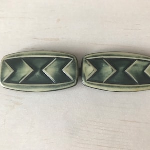 May include: A pair of vintage belt buckles with a geometric design. The buckles are made of a light green plastic with a dark green pattern.