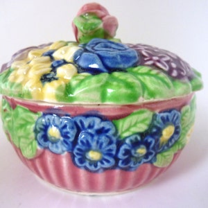 May include: A small, pink ceramic trinket box with a floral design. The lid features a cluster of yellow, blue, and purple flowers with green leaves. The box itself has a pink and white striped pattern with blue and yellow flowers around the bottom.