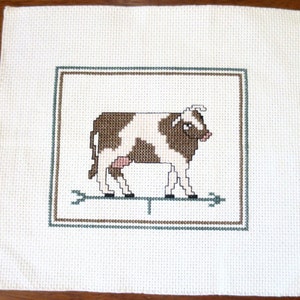 May include: A brown and white cow with a pink udder is embroidered on a white fabric with a green and brown frame.