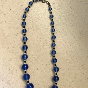 May include: A necklace with blue, black, and clear beads. The necklace features large, round, cobalt blue beads, with smaller black and clear beads. It has a silver-tone clasp. The necklace is displayed against a neutral background.