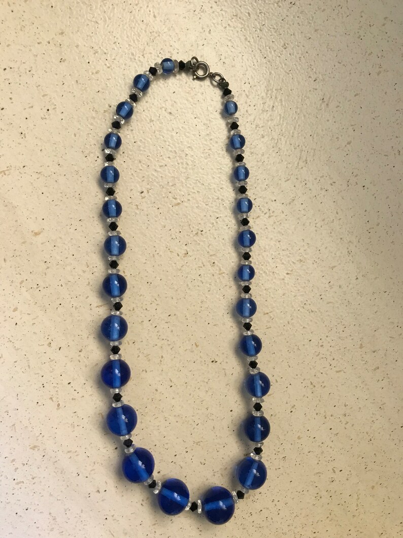 May include: A necklace with blue, black, and clear beads. The necklace features large, round, cobalt blue beads, with smaller black and clear beads. It has a silver-tone clasp. The necklace is displayed against a neutral background.