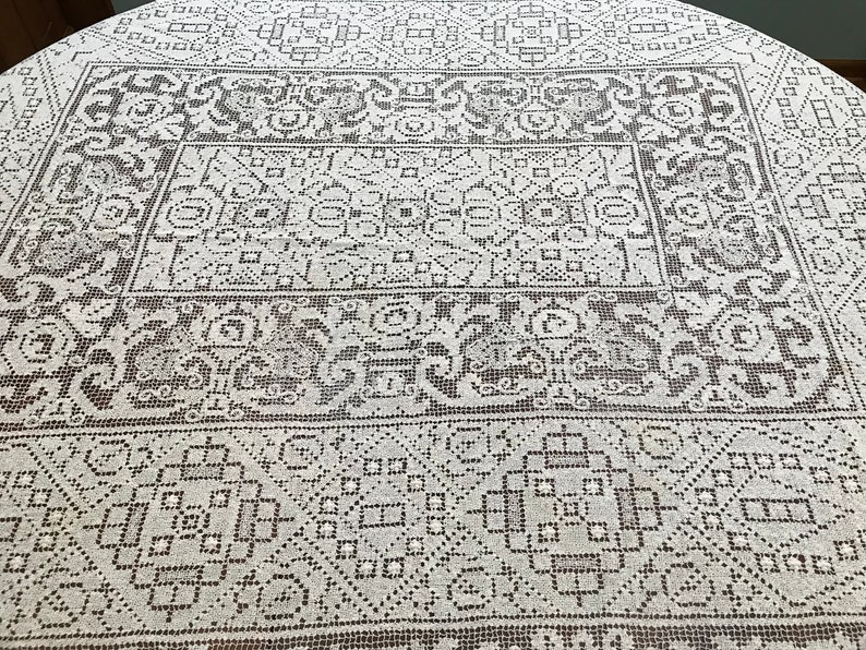 May include: A white tablecloth with a delicate, intricate lace pattern in a dark brown color. The pattern is geometric and symmetrical, with repeating motifs of squares, circles, and flowers.