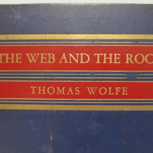 May include: A blue book cover with gold and red accents. The title "THE WEB AND THE ROCK" is printed in gold lettering. The author's name, "THOMAS WOLFE", is printed in gold lettering below the title.