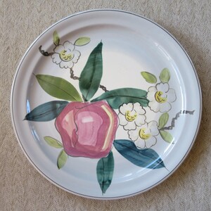 May include: White ceramic plate with a hand-painted floral design. The plate features a large pink apple-like fruit, green and blue leaves, and white flowers with yellow centers. A thin gray line borders the edge of the plate.