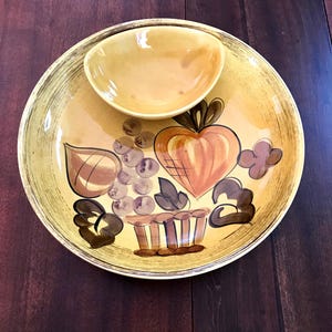 May include: A yellow ceramic serving dish with a smaller bowl attached. The dish features a painted design of fruit in a basket, including a heart-shaped apple, grapes, and leaves.