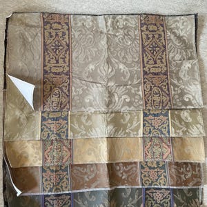 May include: A folded fabric sample featuring a variety of patterned sections. The top section is a light beige with a floral design and a dark purple border. Below are sections in shades of beige, brown, and green, each with a different pattern. The fabric has a textured appearance.