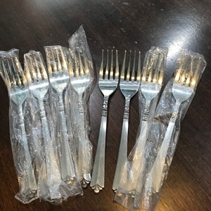 May include: Several silver forks, some in individual clear plastic wrappers. The forks have a decorative pattern on the handles and are arranged on a dark brown wooden surface. The fork tines are clearly visible.