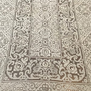 May include: A white crocheted tablecloth with a delicate, intricate pattern. The pattern features a repeating design of squares, circles, and floral motifs.