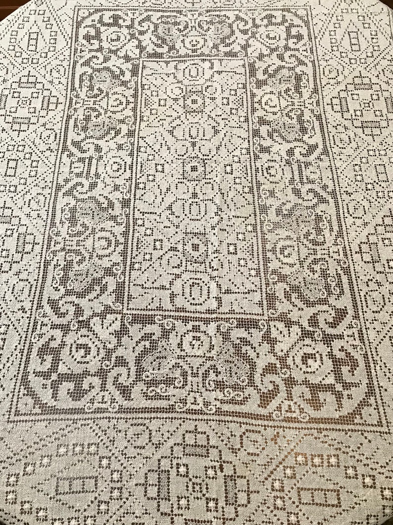 May include: A white crocheted tablecloth with a delicate, intricate pattern. The pattern features a repeating design of squares, circles, and floral motifs.