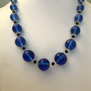May include: A blue beaded necklace with large, round, translucent beads. The necklace features smaller black and clear beads between the larger blue beads. The necklace is displayed on a white mannequin.