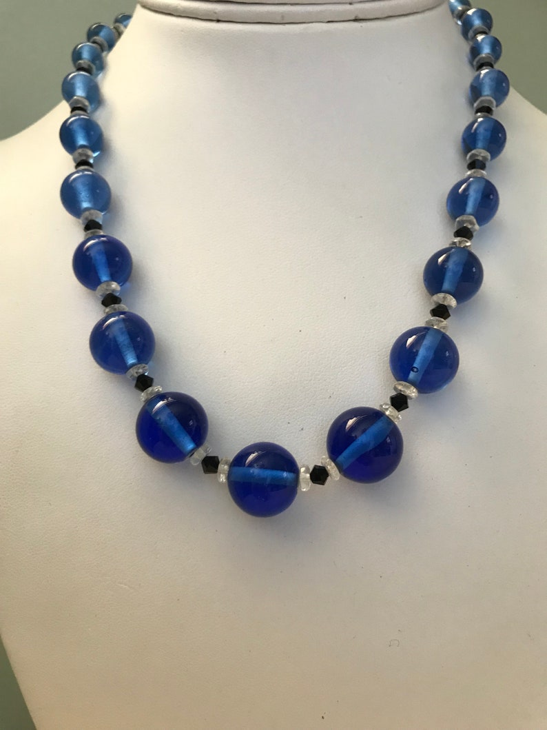 May include: A blue beaded necklace with large, round, translucent beads. The necklace features smaller black and clear beads between the larger blue beads. The necklace is displayed on a white mannequin.