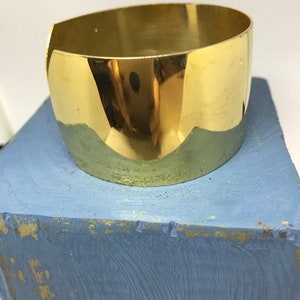 May include: A shiny gold metal cuff bracelet. The bracelet is wide and has a rounded shape.