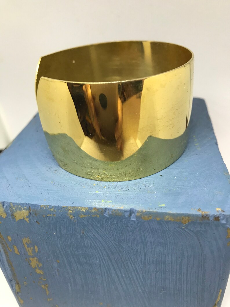 May include: A shiny gold metal cuff bracelet. The bracelet is wide and has a rounded shape.