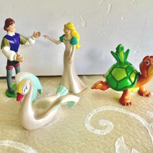 Peut inclure: Ensemble de figurines en plastique vintage représentant un prince en bleu et vert, une princesse en robe blanche, un cygne blanc avec des accents bleus et une tortue verte et orange.