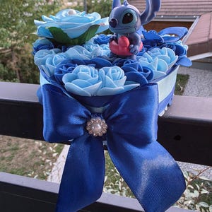Peut inclure: Composition florale bleue avec une figurine Stitch tenant un cœur rouge. L'arrangement comprend des roses bleues de différentes nuances et un grand nœud en satin bleu avec une perle. Environ 15 cm de diamètre.