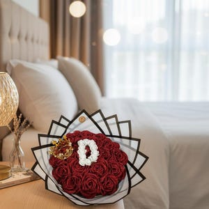Könnte beinhalten: Ein Strauß roter Rosen mit einem weißen Buchstaben "D" in der Mitte, akzentuiert mit einer goldenen Krone. Der Strauß ist in einem schwarz-weißen geometrischen Design verpackt. Die Anordnung befindet sich auf einem Nachttisch aus Holz.