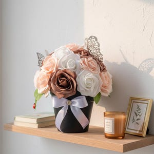 Peut inclure: Un pot de fleurs noir rempli de fausses roses blanches, pêche et marron, orné de papillons argentés et d'un ruban blanc. Le pot est posé sur une étagère en bois avec une bougie et une œuvre encadrée.