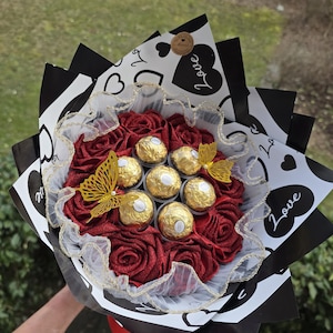 Pode incluir: Um buquê com rosas vermelhas de tecido e chocolates dourados, acentuado com decorações de borboletas douradas. O buquê é embrulhado em papel preto e branco com desenhos de corações e a palavra "Love".