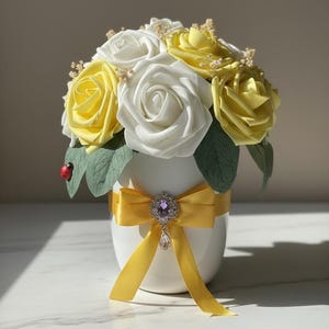 Op de afbeelding: Een witte keramische vaas bevat een boeket gele en witte rozen. Een geel lint met een juweel versiert de vaas. Een klein lieveheersbeestje zit op een groen blad.