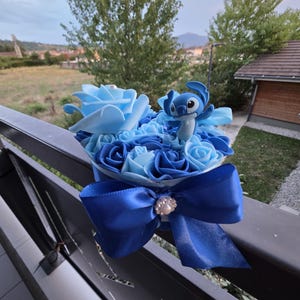 Peut inclure: Un bouquet de roses bleues avec une figurine Stitch, noué avec un ruban de satin bleu foncé. Le bouquet présente différentes nuances de bleu, du clair au foncé, et est orné d'une décoration en forme de perle au centre du nœud.