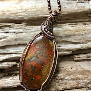 May include: A handcrafted pendant necklace featuring a teardrop-shaped stone with red, orange, yellow, and brown patterns. The stone is encased in copper wire with intricate wrapping details and hangs from a copper ball chain.