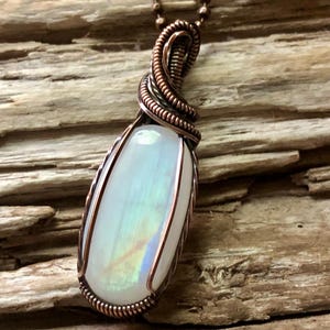 May include: An oval-shaped white moonstone pendant wrapped in copper wire, suspended from a matching copper ball chain. The stone displays a subtle iridescent sheen, and the metal wire is coiled tightly around the top of the gemstone.