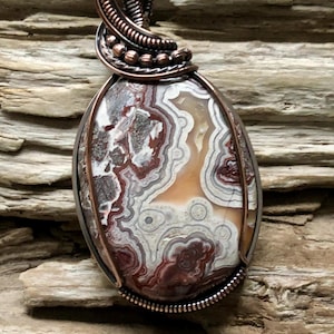 May include: Oval pendant featuring a patterned stone in shades of brown, white, and tan, encased in a copper wire wrap. The pendant hangs from a copper chain, showcasing a unique, handcrafted design. The stone has a polished surface.