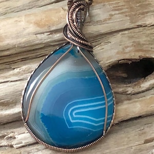 Op de afbeelding: Een druppelvormige hanger met een blauwe agaatsteen met witte banden, ingekapseld in een koperdraadframe. De hanger hangt aan een koperkleurige ketting en toont een handgemaakt ontwerp van natuursteen.