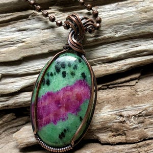 May include: Oval pendant necklace featuring a green and magenta stone with copper wire wrapping. The stone has black speckles and is suspended from a copper ball chain. The pendant is approximately 1.5 inches long.