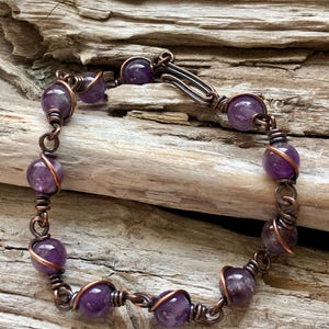 May include: A purple amethyst gemstone bracelet with copper wire wrapping each bead. The bracelet has a silver clasp.