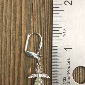 Puede incluir: Un pendiente de plata con un dije de &aacute;ngel. El cuerpo del &aacute;ngel es de cristal transparente y sus alas son de plata. El pendiente mide aproximadamente 2,5 cm de largo y se muestra junto a una regla.