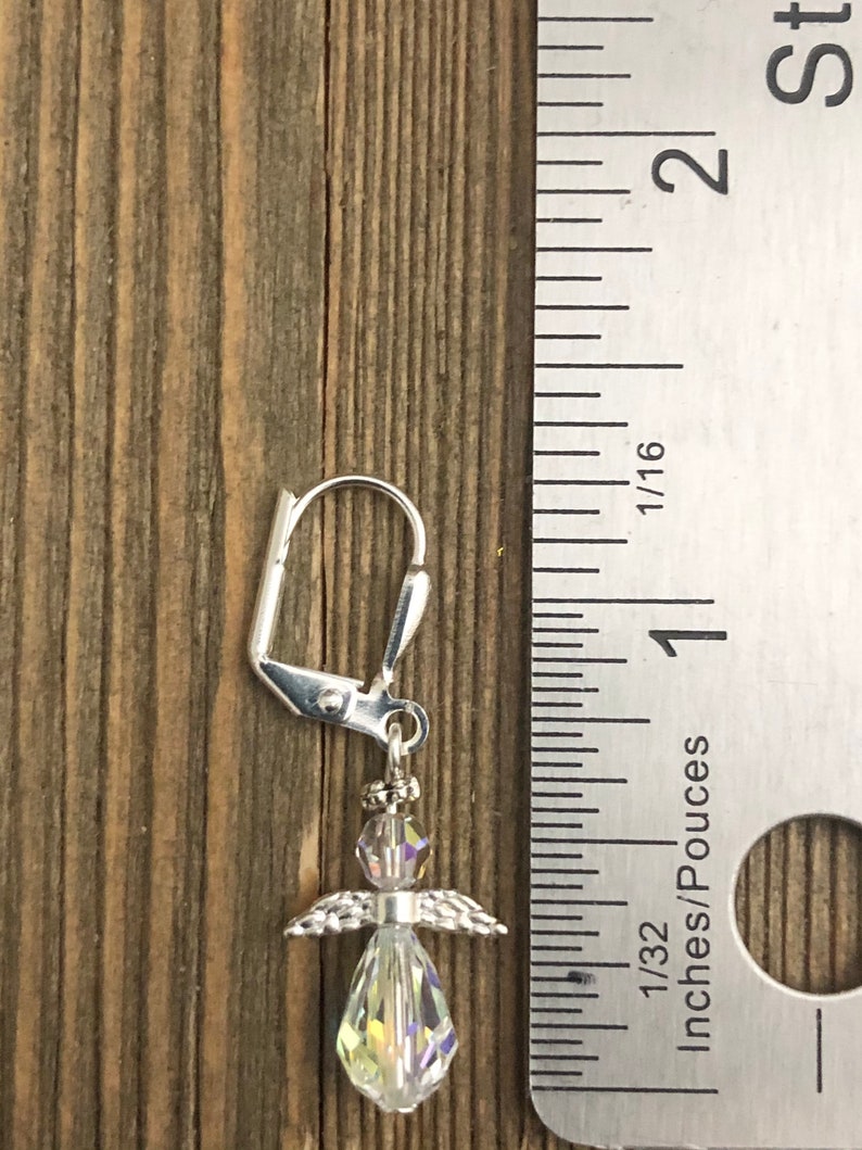 Puede incluir: Un pendiente de plata con un dije de &aacute;ngel. El cuerpo del &aacute;ngel es de cristal transparente y sus alas son de plata. El pendiente mide aproximadamente 2,5 cm de largo y se muestra junto a una regla.