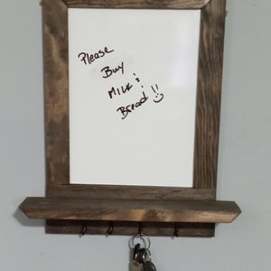 May include: A rustic wooden framed whiteboard with a shelf and hooks underneath. The whiteboard has a handwritten grocery list that says "Please Buy Milk & Bread" with a smiley face. There are keys hanging on the hooks.