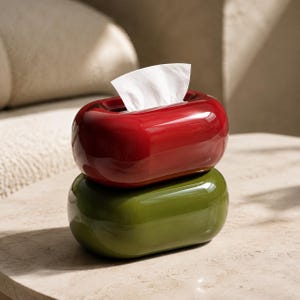 Puede incluir: Una pila de dos cajas para pañuelos de forma ovalada con acabado brillante. La caja superior es de un color rojo vibrante, mientras que la inferior es de un tono verde oliva intenso. Un pañuelo blanco sobresale por la abertura central superior.